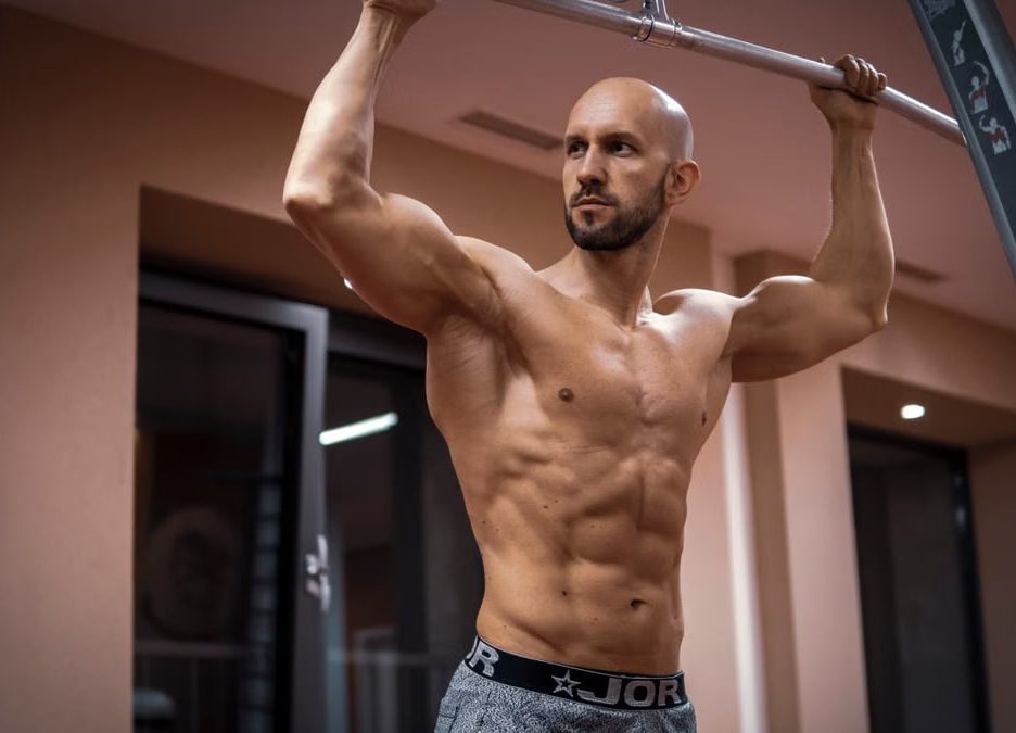 A bald, muscular man performing exercise on gym equipment indoors.